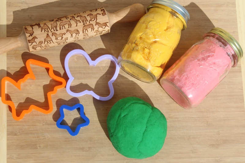Ball of green gluten free playdoh on a cutting board with two jars of yellow and pink playdoh, cookie cutters, and a rolling pin