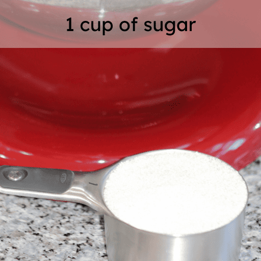 1 cup of sugar in a stainless steal measuring cup sitting on the counter in front of a red kitchenaid stand mixer. Ingredient and measurement is listed at the top of the image. 