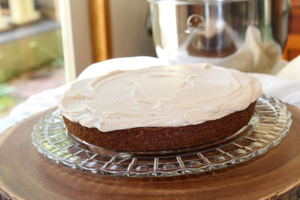 One layer of apple spice cake on the crystal cake plater with cinnamon buttercream frosting smoothed over the top. The platter is on a wood tree slice surrounded by white linen with the mixing bowl of frosting in the background.