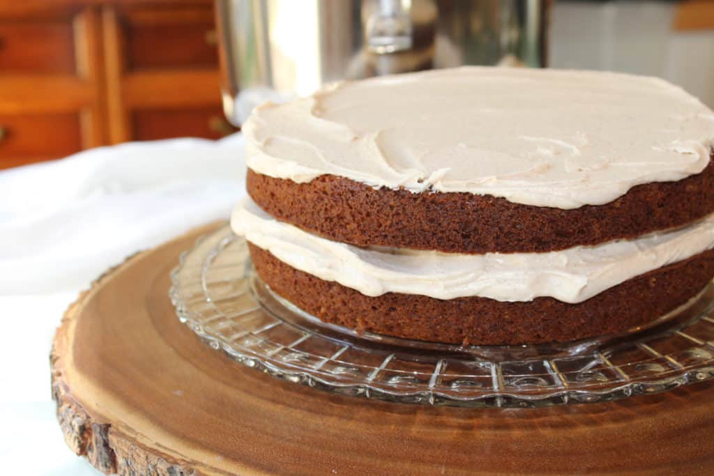 Two layers of apple spice cake on a crystal cake plater each topped with cinnamon buttercream frosting smoothed over the top. The platter is on a wood tree slice surrounded by white linen with the mixing bowl of frosting in the background.