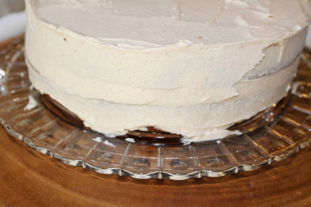 Apple spice cake with two layers with icing on the top and sides. It is sitting on a crystal platter on a wood tree slice.
