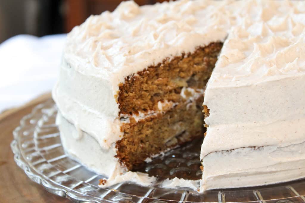 Apple spice cake on a crystal cake platter. One slice has been taken out of the cake. The cake is golden brown with apple chunks. The icing is a creamy white with flecks of cinnamon. The platter is sitting on a tree slice which is on white linen.