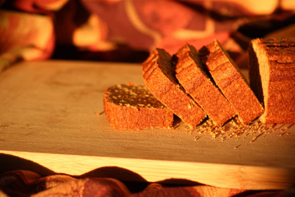 side view of gluten free pumpkin bread with 4 slices leaning over on the cutting board. The rest of the loaf is directly behind the slices. The cutting board is sitting on a gathered warm toned table cloth. The lighting is warm and brings out the amber tones. 