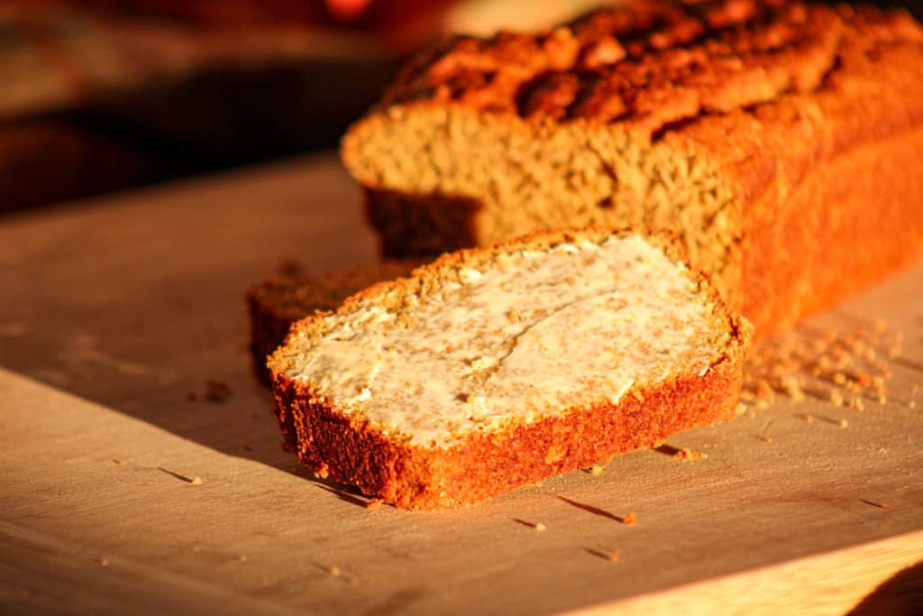 One slice of gluten free pumpkin bread with butter sitting on a cutting board in front of the rest of the loaf.