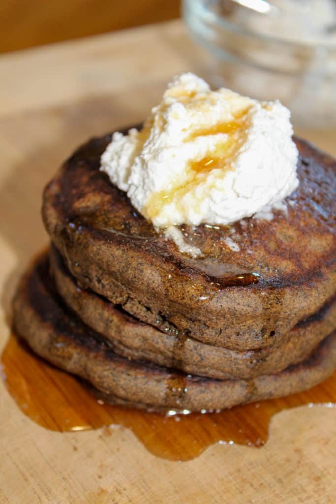 Stack of three pumpkin spice pancakes sitting on a wooden cutting board topped with dairy free whipped topping and maple syrup 