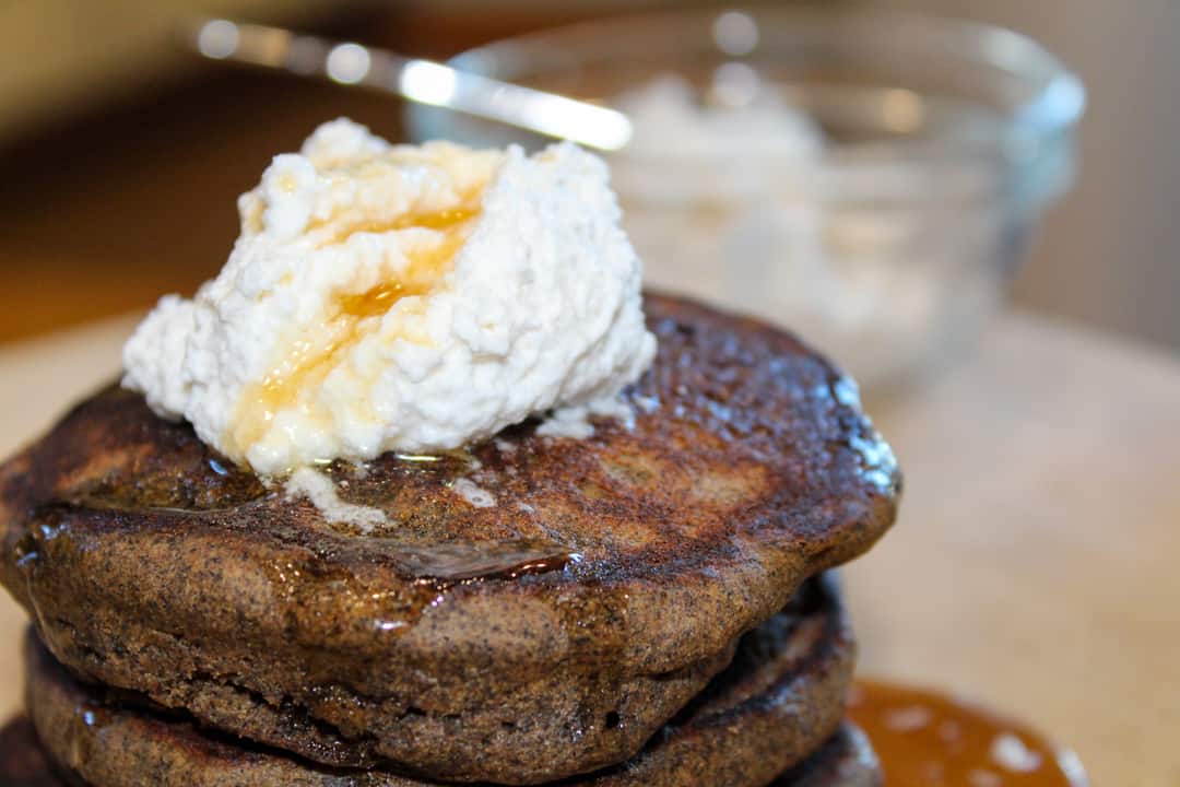 tall stack of pumpkin spice pancakes topped with whipped cream and maple syrup sitting on a wooden cutting board. In the background is a small glass bowl of whipped cream with a silver spoon.