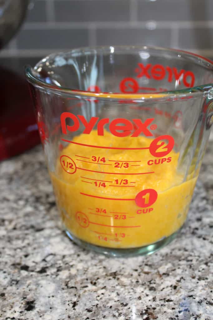 1 cup of pumpkin puree in a glass 2 cup measuring cup. The measuring cup is sitting on the counter in front of a red kitchen aid mixer. 