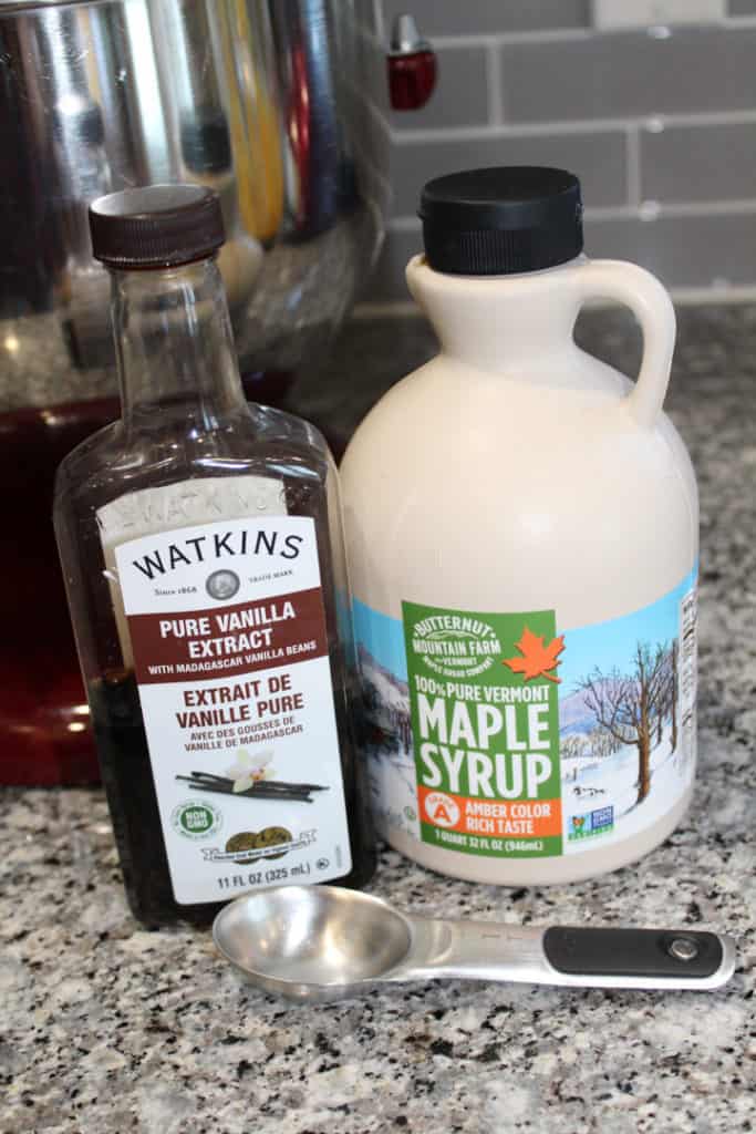 Container of vanilla extract sitting next to a thing of maple syrup on the counter in front of a mixer. A 1 tablespoon measuring spoon is sitting on the counter in front of the containers. 