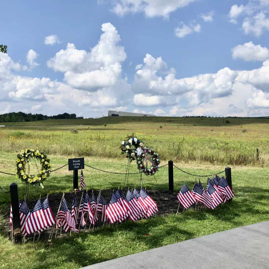 flags and wreaths in the grass next to a sidewalk at the 93 memorial field. behind the wreaths are a few posts, rope, and a sign. 