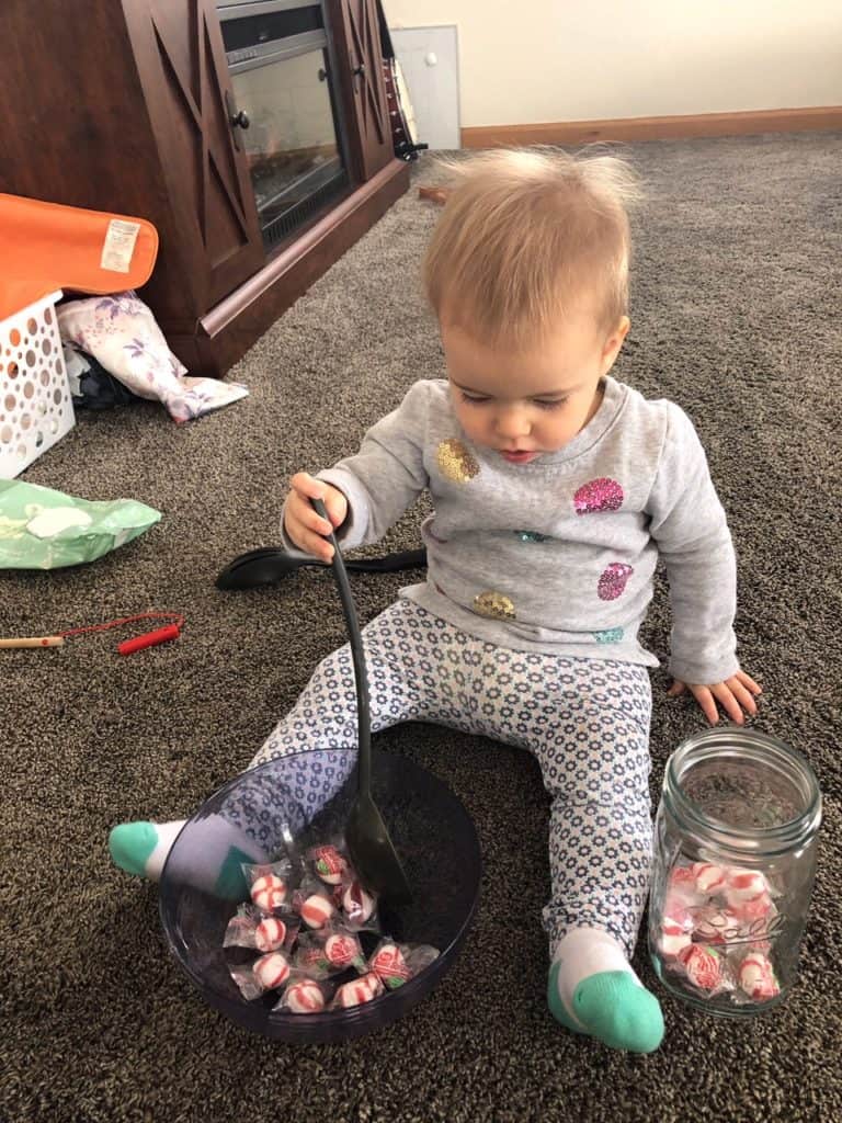 1 year old girl sitting on the floor with a plastic bowl filled with peppermints between her legs. She is holding a ladle in her right hand. To her left is a jar holding a few peppermints. She is scooping the peppermints from the bowl and dumping them in the jar. 