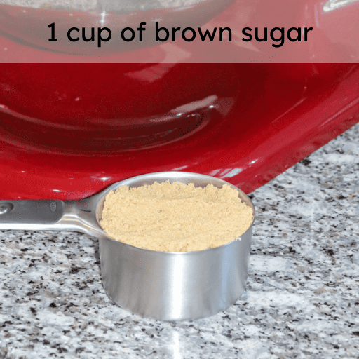 1 cup of brown sugar in a stainless steal measuring cup sitting on the counter in front of a red kitchenaid stand mixer. Ingredient and measurement is listed at the top of the image. 