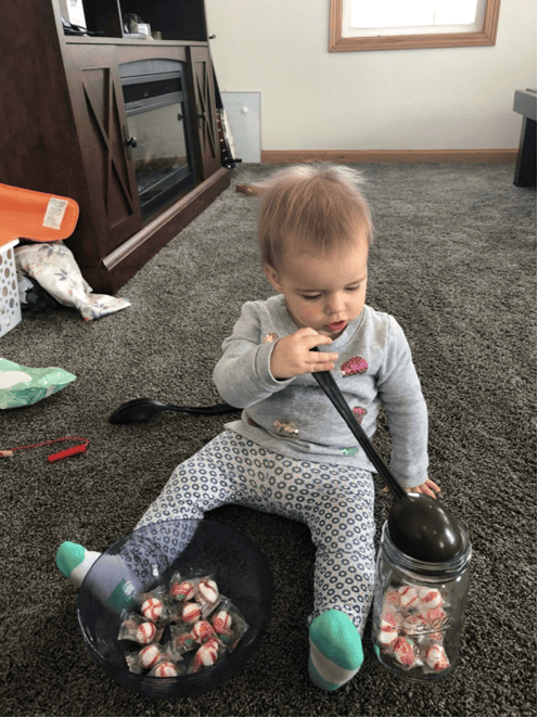 Toddler using eye movement skills to look at the mason jar to her left as she dumps the peppermint candies from her ladle into the jar. 