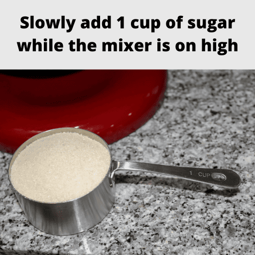 one cup of sugar in a stainless steal measuring cup sitting on the counter in front of a red mixer stand. 