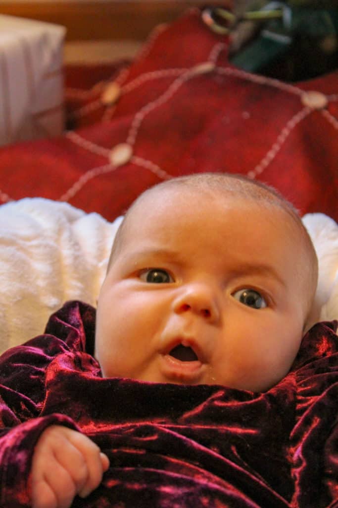 2 month old baby laying propped up on a boppy covered in a white blanket. In the background is a red Christmas tree skirt, a Christmas tree, and a present. The baby's eye movement skills are still developing. Her eyes are not in proper alignment while trying to focus on the camera. 