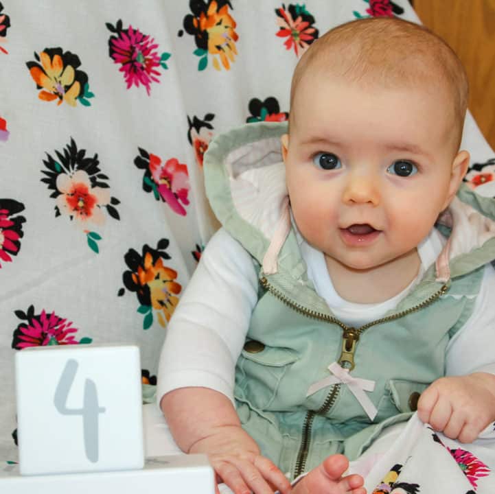 4 month old baby girl in a white long sleeve onesie with a light green vest on. She is sitting in the corner of a rocking chair covered in a flower blanker. There is a white block with a gray number four on it in front of her. The baby is slightly leaning forward grasping the blanket with her left hand looking at the camera with both eyes moving in alignment. 