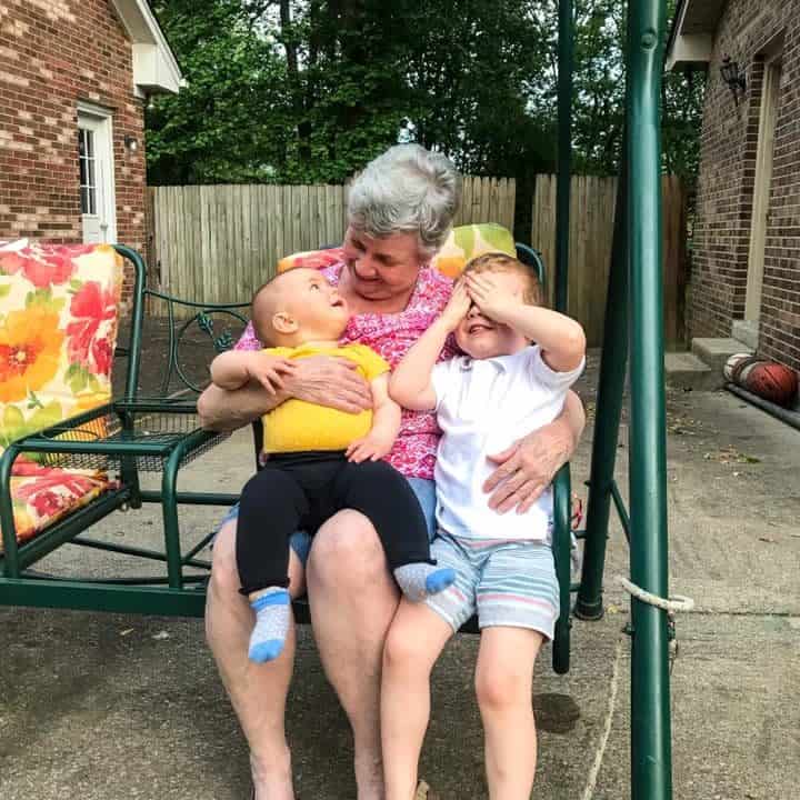 A grandma with a baby and a toddler on her lap outside on a green porch swing.