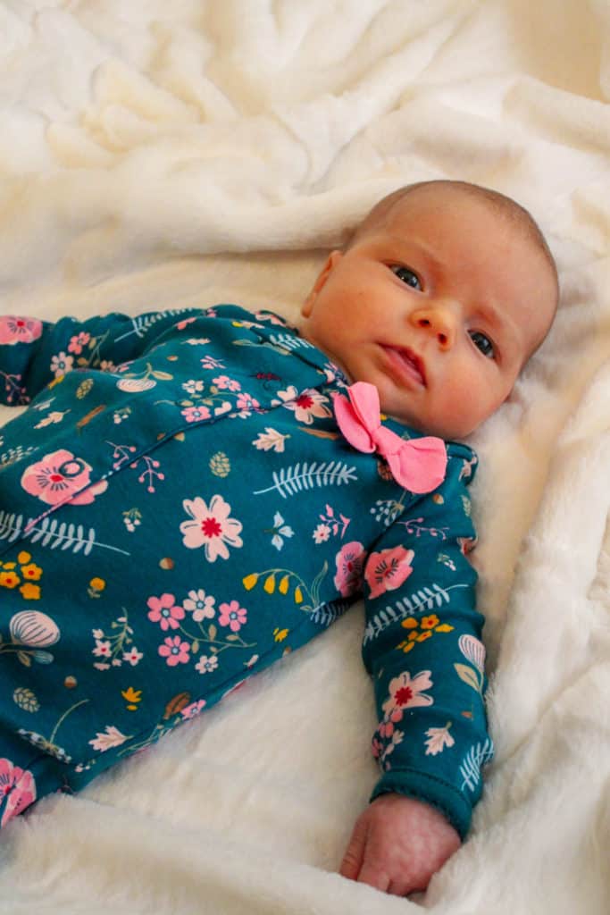 1 1/2 month old baby laying on a white blanket looking at the camera. She is wearing a green outfit covered in flowers that are various shades of pink. The baby's eye movements skills are developing and at this moment her eyes are working together to look at the camera. 