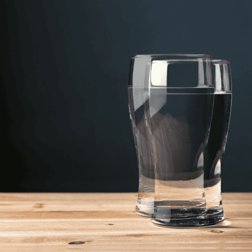 A glass water cup sitting on a wooden table in front of a blackish blue background. There is a second blurry image of the same cup overlaid over the main cup to show what someone with a vision difficulty like double vision would see. 