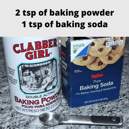 A container of baking powder on the left and a container of baking soda on the right with a measuring spoon sitting on the counter in front of them.