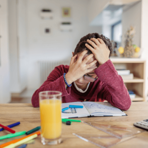 Middle school aged kid sitting at the table working on math homework. Resting hand on his hands covering one eye. A glass of orange juice is in front of him and there are an array of colored pencils on the table. There are protractors on the table