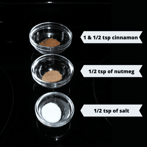 Three small glass bowls sitting on a black surface in a vertical line in descending size. The top bowl has cinnamon, the middle has nutmeg, and the bottom has salt.