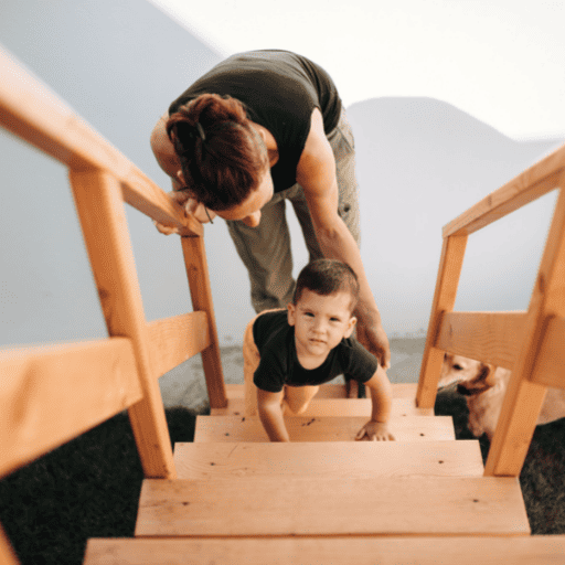 toddler aged boy crawling up the stairs with his mother behind him. He is squinting and looking up the staircase. The stars are wooden and very airy and open. There is a wooden railing on both sides. The mom is bent over with one hand on the railing and one hand protectively reaching down.