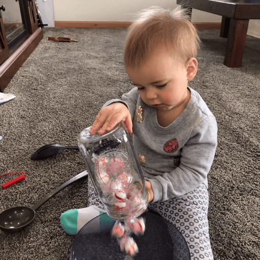 1 year old girl using eye movement skills to pour a glass jar full of soft peppermints in a plastic bowl sitting on the floor in front of her. The bowl is between her legs and there are two ladles/spoons to her right. 