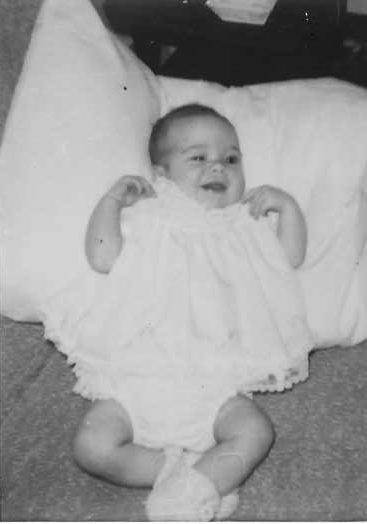 Infant laying against a pillow on the floor. Image shows poor development of baby's eye movements. One eye is clearly open and the other is half closed as she focuses on something outside of the photo.