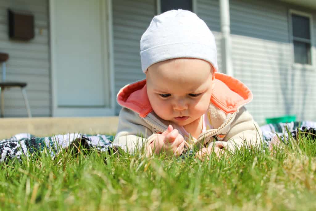 baby girl wearing a hoodie and hat laying on her belly outside on a blanket. She is in front of a gray house. She is at the edge of the blanket holding a blade of grass with one hand. She is intently inspecting the grass.