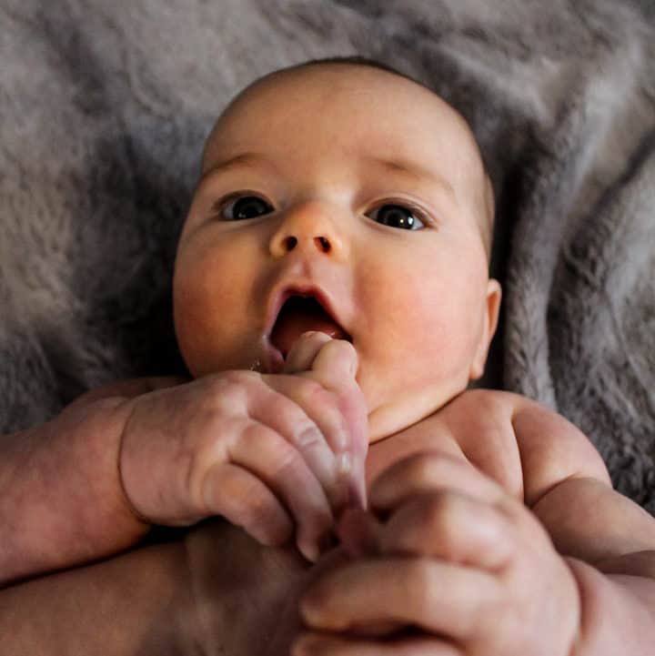 3 month old laying on a gray blanket. She is hold each of her feet with one hand and bringing them to her mouth. Her eyes are looking up at the camera above her. 