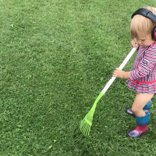 Toddler girl wearing a red, white, and blue long sleeve one piece swimsuit, ear muffs, and rain boots raking grass clippings.