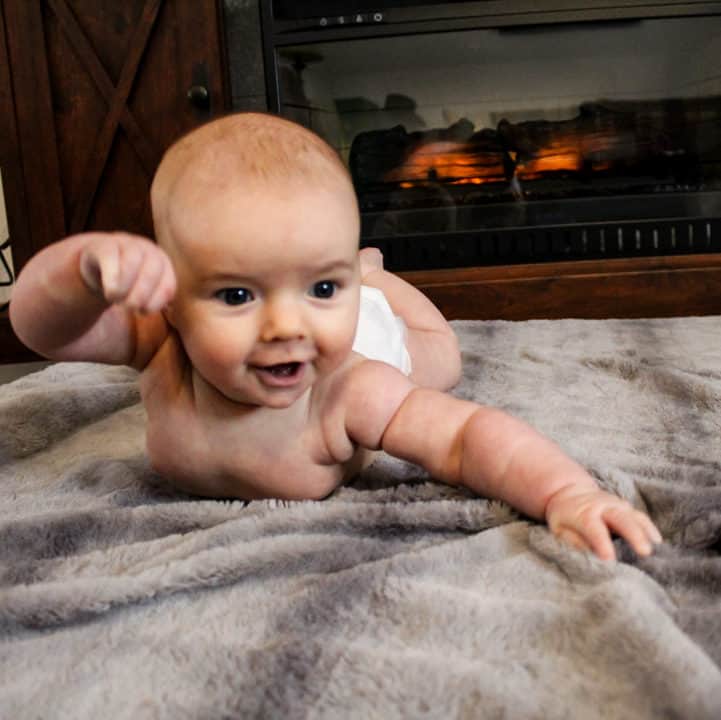 4 month old on her tummy on a gray blanket in front of a fake fireplace tv stand. She is intently looking at an object and moving her arms in an attempt to move closer to it. The baby's eye movements are allowing her eyes to work together and focus on the object. 