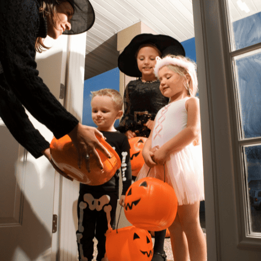 three kids dressed up trick or treating at dusk. There are standing in a doorway. The homeowner is a woman wearing a black witch hat holding an orange bowl of candy. 