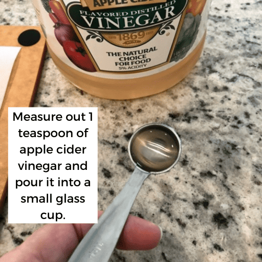 hand holding 1 teaspoon of apple cider vinegar above the countertop with the apple cider vinegar bottle in the background.