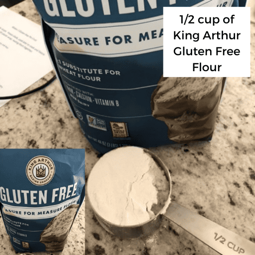 A 1/2 cup measuring cup filled with king arthur gluten free flour sitting on the countertop. The bag of flour is sitting directly behind the measuring cup. 