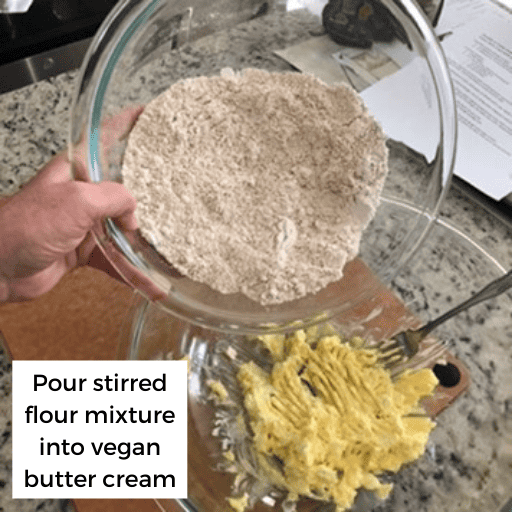 Hand holding a glass bowl with a flour mixture tilted above a bowl with a vegan butter cream mixture. The second bowl is sitting on a cutting board on the counter. 