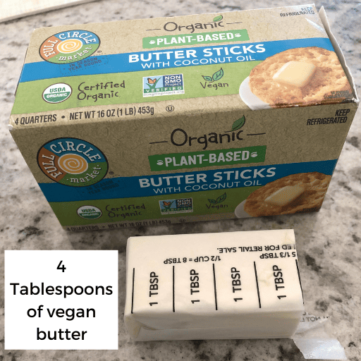 4 tablespoons of vegan stick butter sitting on the countertop. The butter box is directly behind the stick of butter.