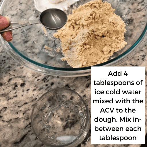 large glass bowl with the gluten and dairy free pie crust dough in the top half of the image. One tablespoon of ice cold water is being held above the bowl. In the lower half of the image is a small glass cup with water and an ice cube in it.