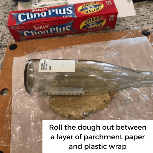 gluten and dairy free pie crust dough being rolled out between a sheet of parchment paper and plastic wrap on a cutting board. A red box of plastic wrap is sitting on the counter above the cutting board. A glass bottle is sitting on top of the plastic wrap to roll out the dough. 
