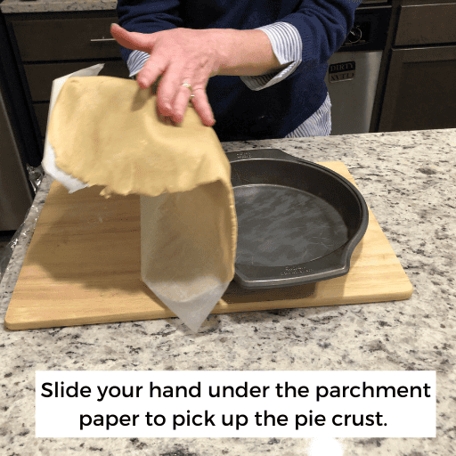 person holding pie crust on wax paper between their hands above a pie pan. The pie pan is sitting on a cutting board on a kitchen island. 