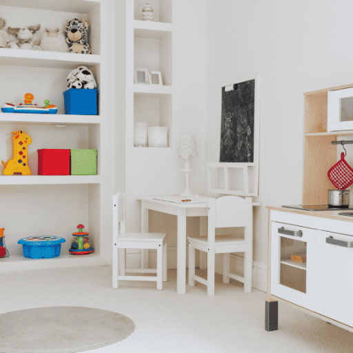 Playroom with open shelves on the back wall. Toys are accessible and not in bins. There is a white toddler table with two chairs in the corner. On the wall on the right there is a play kitchen