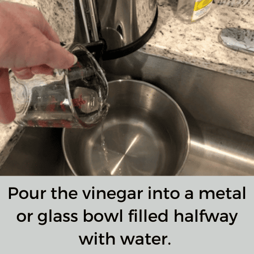 Glass measuring cup of apple cider vinegar being poured into a metal bowl with water in the sink. 
