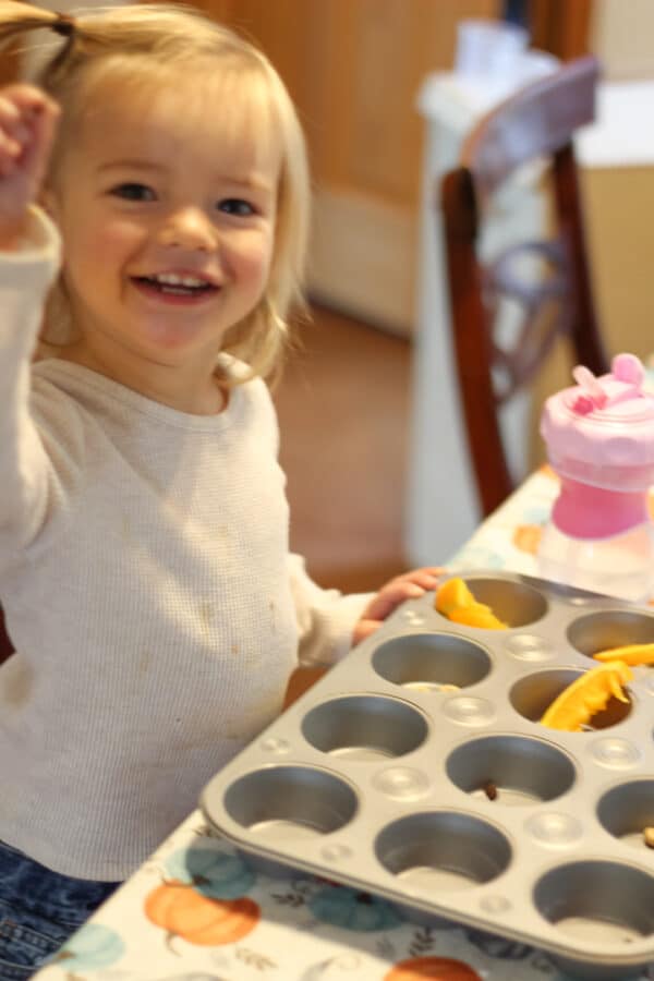 Little girl with blond hair sitting at a kitchen table. She has a cookie sheet in front of her with snacks. She is smiling at the camera holding a cheerio up.