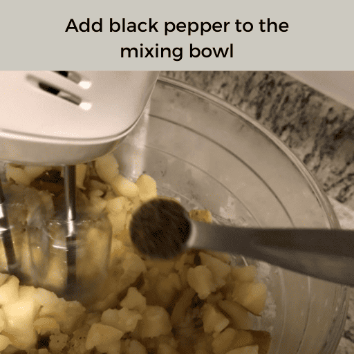 1/4 tsp measuring spoon of black pepper being dumped into a stand mixer's mixing bowl of creamy mashed potatoes.