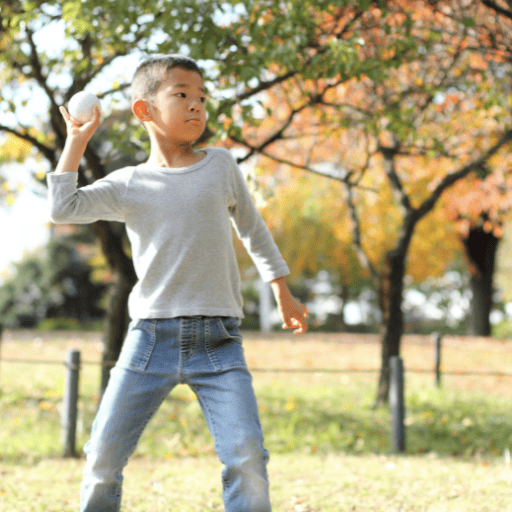 preschool aged boy wearing long sleeve and jeans playing catch outside. It is fall and the leaves on the trees behind him are changing color. He is standing with his left foot forward and his right arm back behind his head about to toss the ball