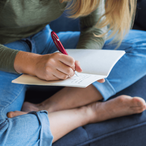 Woman wearing a green long sleeve shirt and a pair of blue jeans that are rolled at the end. She is sitting crossed legged on a blue couch writing in a journal with a red pen. 