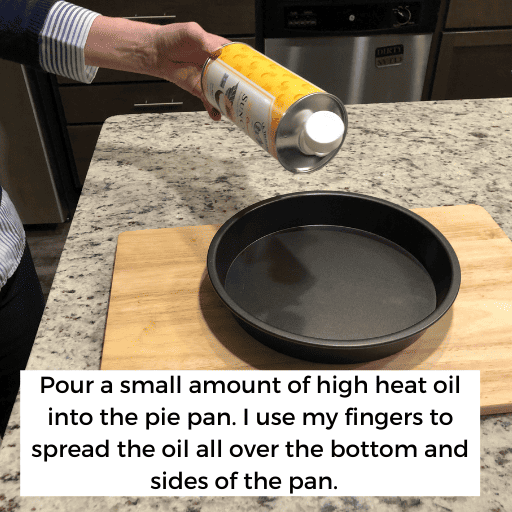 hand holding a bottle of high heat cooking oil above a pie pan. The cake pan is sitting on top of a cutting board on a kitchen island. 