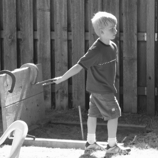 black and white picture of a little boy playing horseshoes. He is wearing a t-shirt, shorts, sneakers, and ankle socks. He has light colored hair. His right arm is stretched out behind him as he prepares to toss the horseshoe. His right foot is in front of the left foot. In the background is a tall wooden fence. Horseshoes is a visual-motor activity.