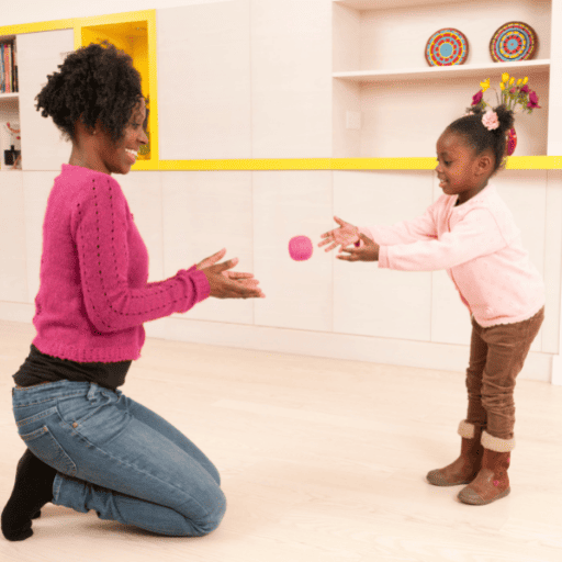 a mother and daughter playing catch in the house with a small pink ball. Playing catch is a visual processing activity. The mom is kneeling. The girl is tossing the ball to her mom.