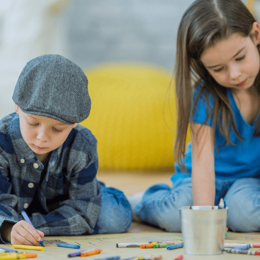 two preschool aged kids sitting on the floor coloring with crayons on a large brown piece of paper. A boy wearing a hat is on the left and he is leaning forward on his forearms. A girl is on the left and she is reaching down with her right hand to color. Multiple crayons are on the paper and in a tin cup in front of them. 